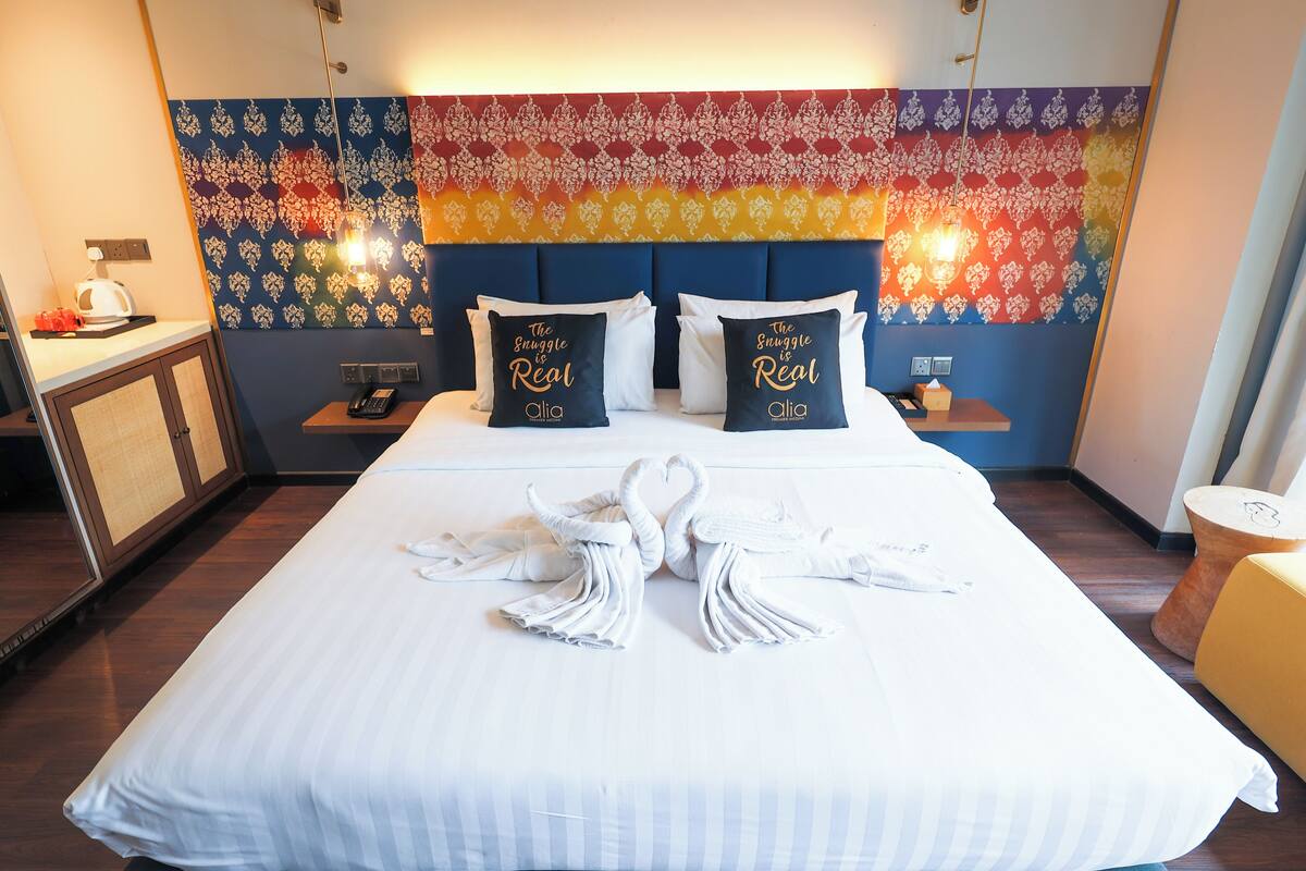 A hotel room with a neatly made bed, two pillows with text, a tapestry on the wall, and a small table with a lamp.