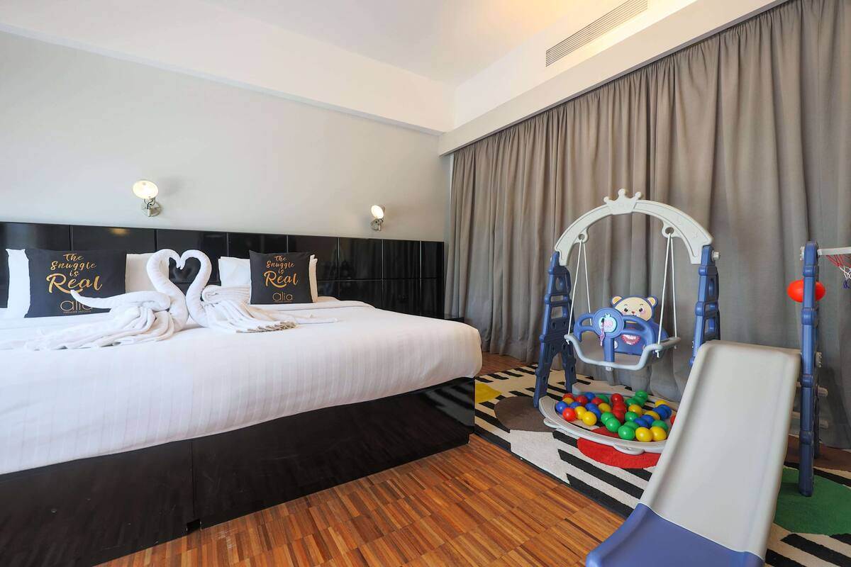 A hotel room with a bed, a headboard with pillows, a wall-mounted lamp, a window with curtains, a play area with a slide and balls, and a small table.