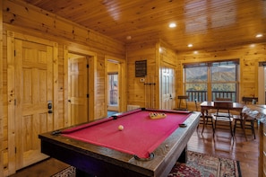 A wooden cabin with a pool table, a dining area, and large windows offering mountain views.