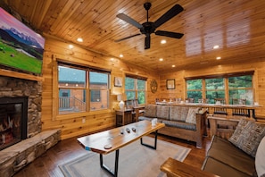 A cozy cabin interior with a stone fireplace, wooden walls, a large TV, a ceiling fan, and a dining area with a view of the outside.