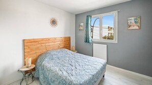 A bedroom with a wooden headboard, a bed with a patterned bedspread, a small bedside table with a lamp, a window with curtains, and two framed pictures on the wall. - Hyères T2 5mn plage Almanarre CLiM/PK/WiFi (HYERES)
