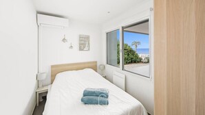 Een slaapkamer met een bed, een nachtkastje, een lamp, een raam met uitzicht op het strand en een wandgemonteerde airconditioner.