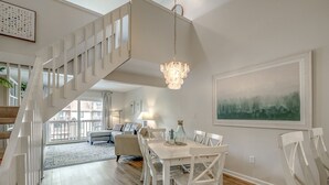 A spacious living area with a white staircase, a dining table with chairs, a chandelier, and a large window with a view.
