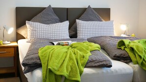 Un lit bien fait, avec des oreillers à carreaux gris et blancs, une serviette verte et un magazine posés sur le lit. Deux lampes de chevet sont placées de chaque côté du lit.