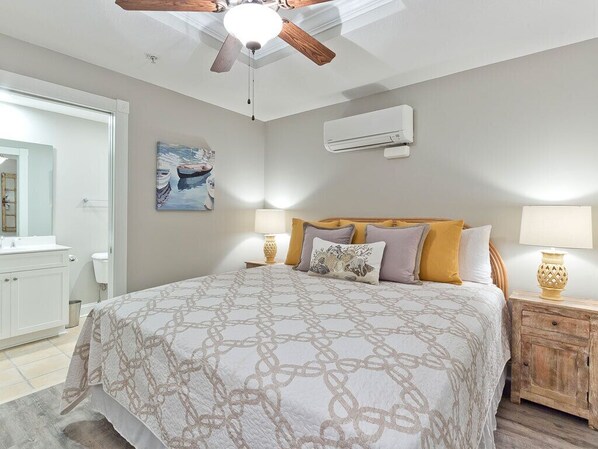 A bedroom with a bed, bedside lamps, a ceiling fan, an air conditioning unit, and a painting on the wall.