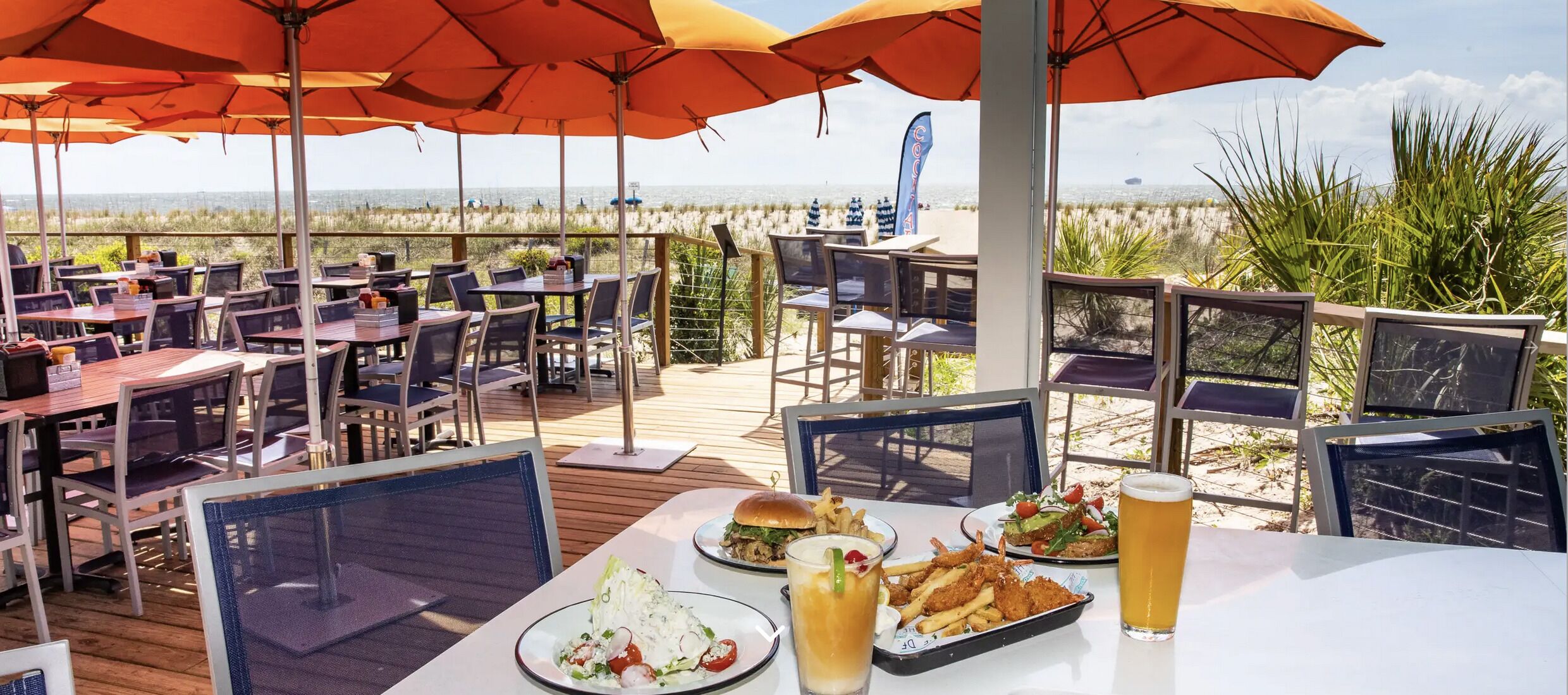 An outdoor dining area with tables and chairs, umbrellas, and a view of the beach.