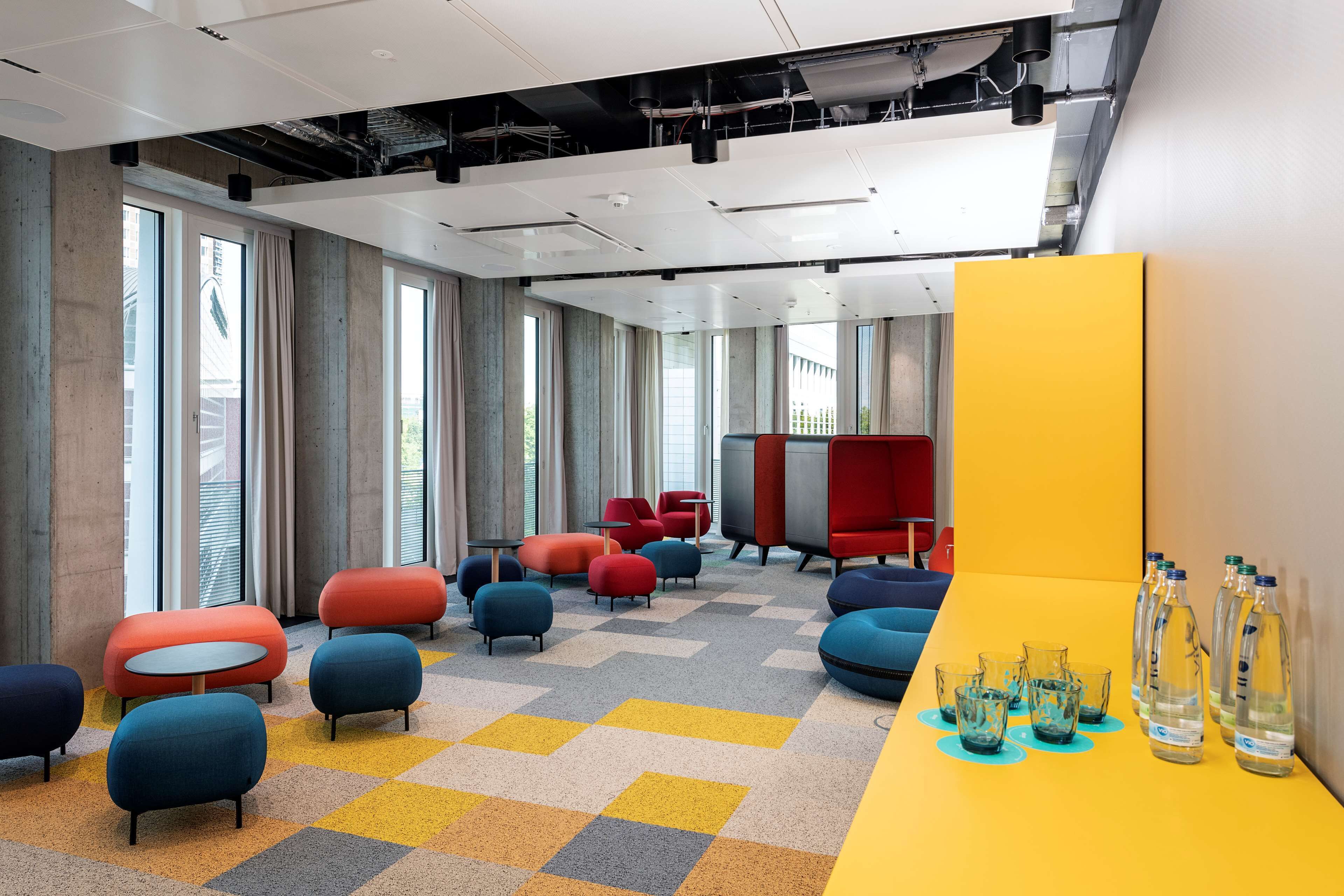 Uma área de estar moderna com assentos coloridos, grandes janelas e uma mesa amarela com garrafas de água.