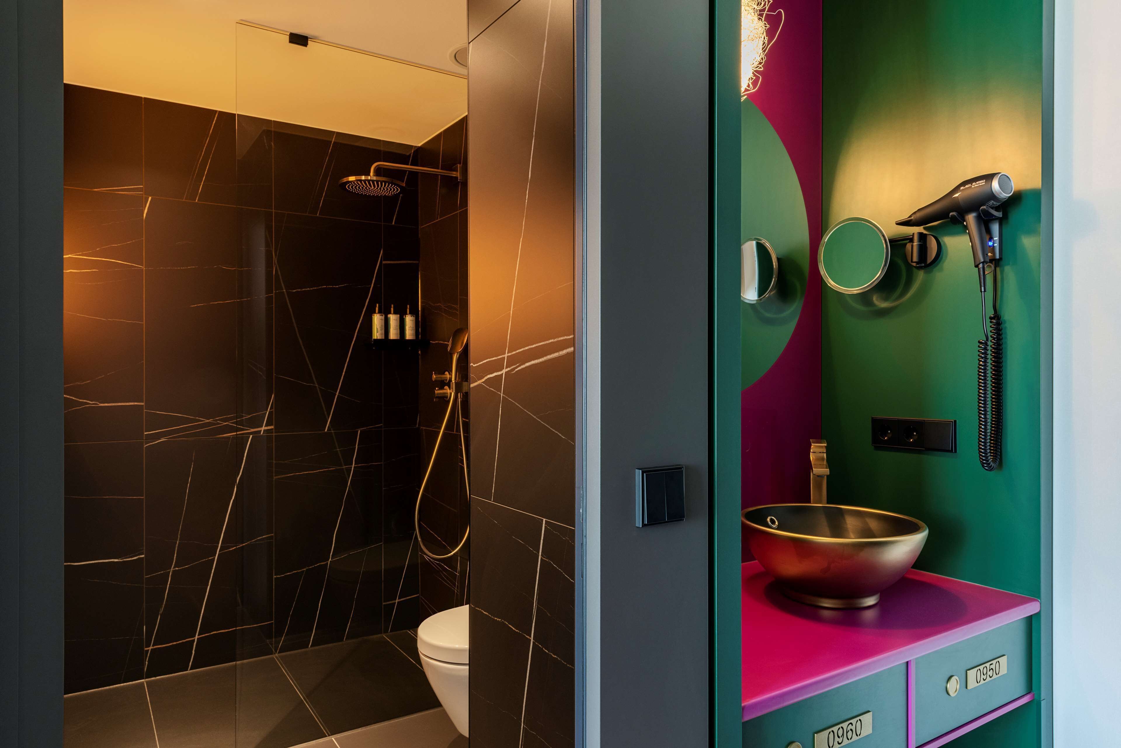 A modern bathroom with a black marble shower, a white toilet, and a vanity with a pink countertop and a gold bowl sink.
