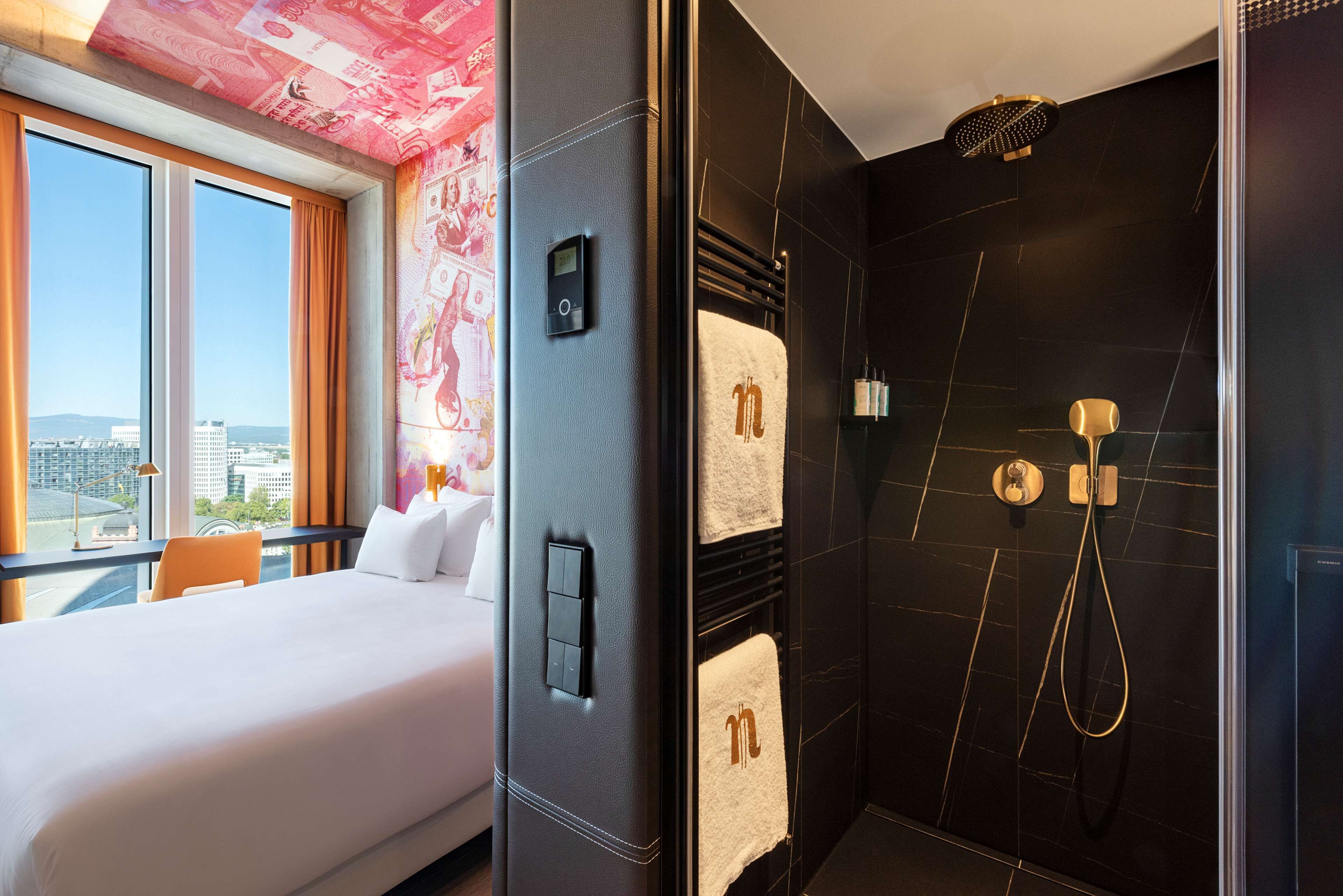 A hotel room with a large bed, a view of the city, a modern shower, and a control panel on the wall.
