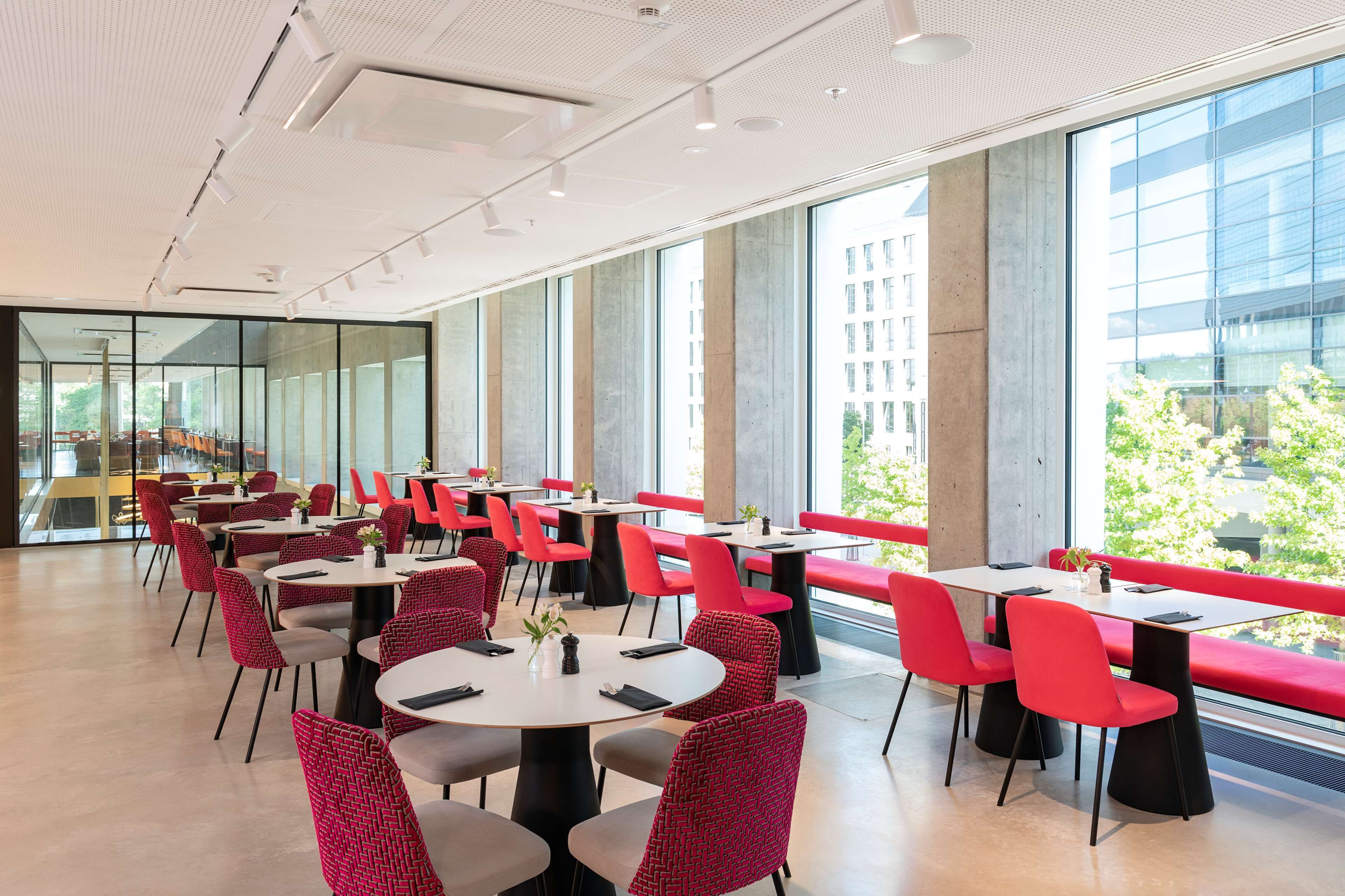 A modern dining area with round tables and red chairs, large windows, and a view of greenery outside.