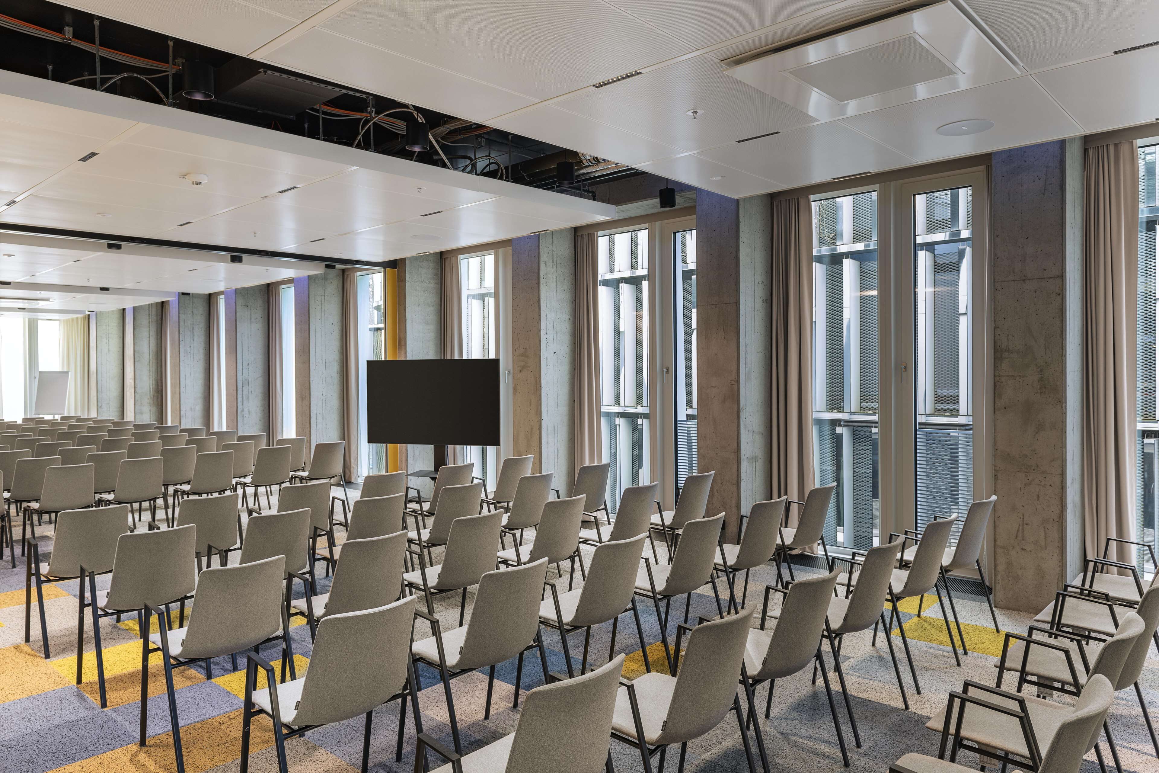 Ein Konferenzraum mit Stuhlreihen, einer großen Leinwand und hohen Fenstern mit Vorhängen.
