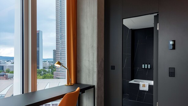 A modern room with a desk, chair, and a view of tall buildings. - nhow Frankfurt (Frankfurt)