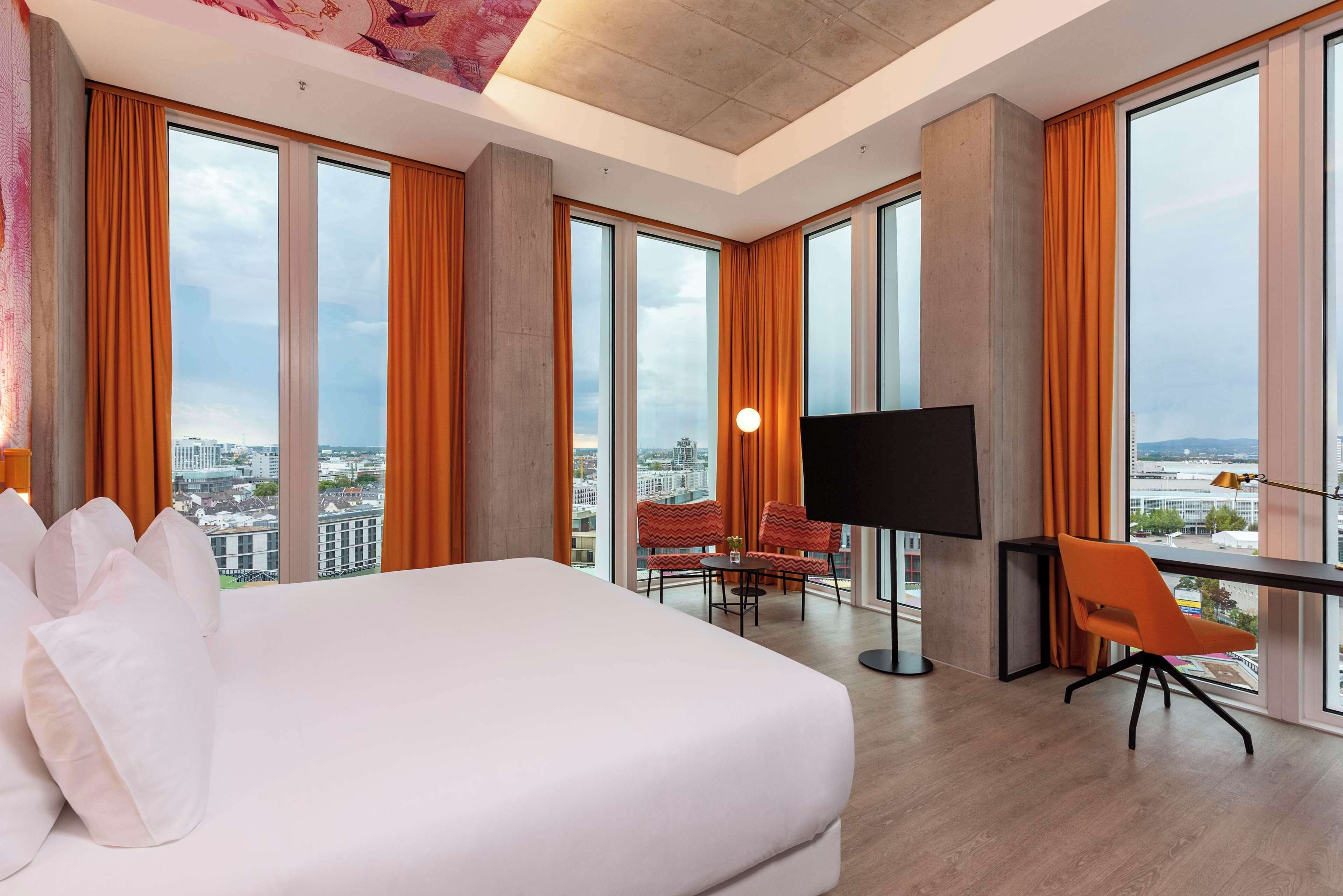 A hotel room with a large bed, a TV, a desk, and a view of the city.