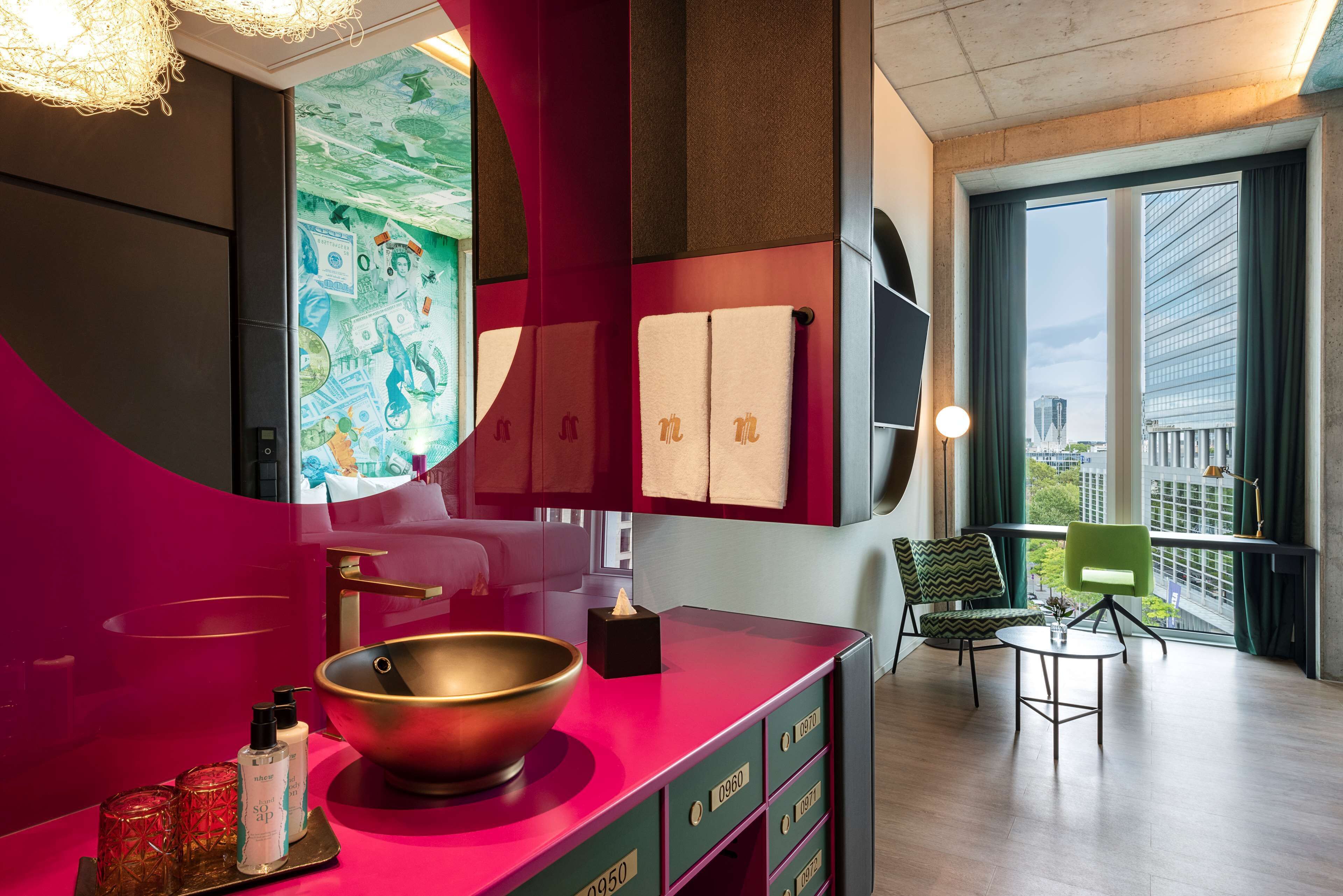 A modern bathroom with a large mirror, a bowl sink, and a vanity with drawers. There is a window with a view of buildings and a chair in the corner.