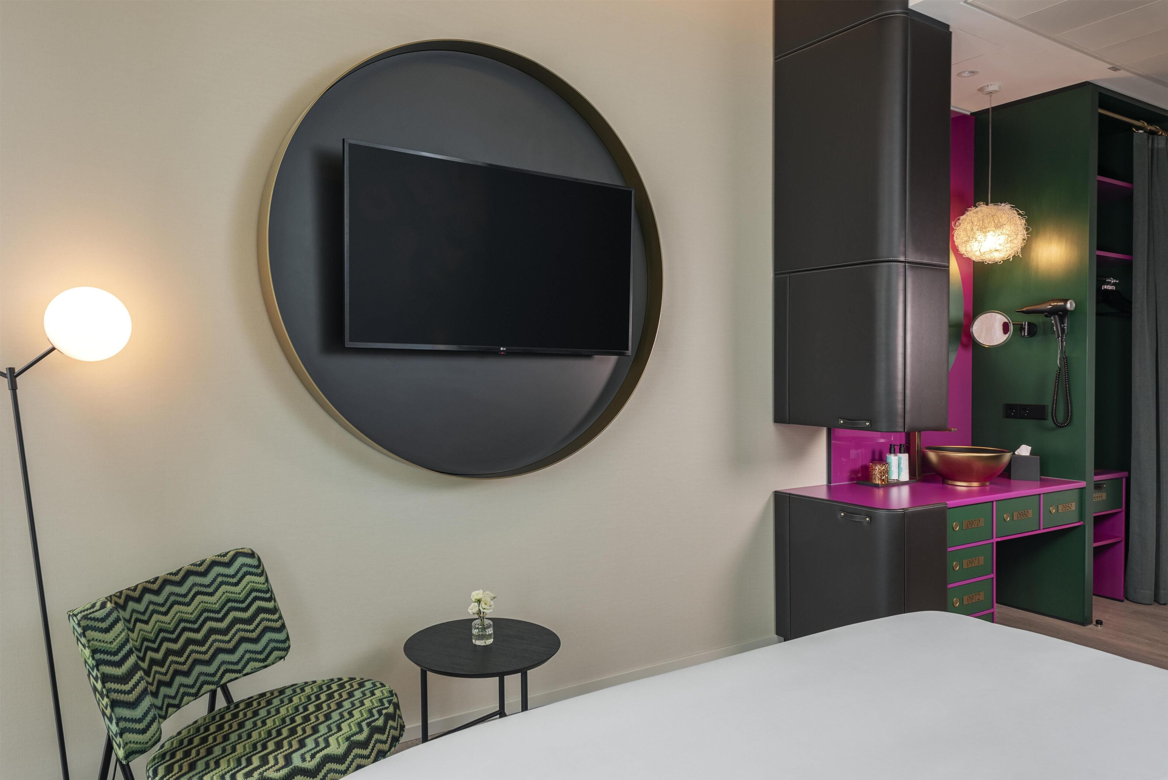 Ein modernes Schlafzimmer mit einem großen Bett, einem runden Wand-Fernseher, einem kleinen Nachttisch mit einer Vase, einem Sessel und einer Kommode mit Spiegel und Fön.
