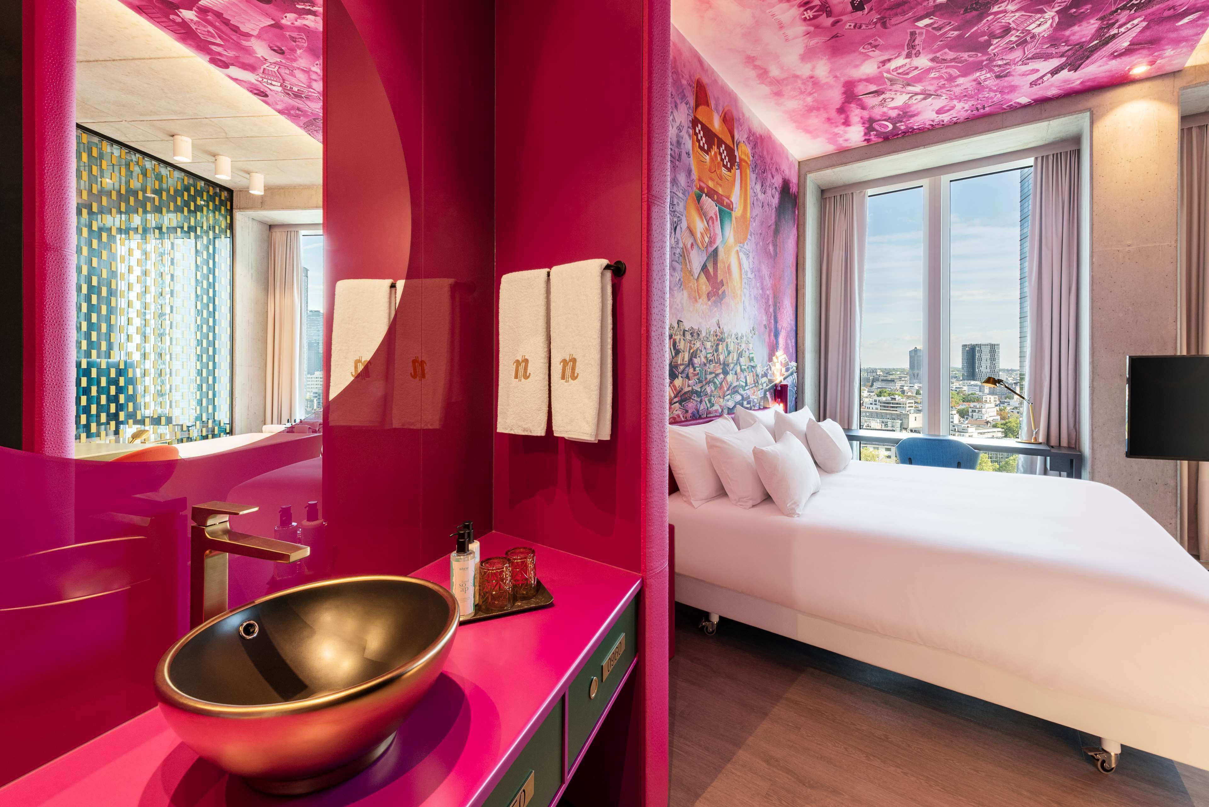 A modern hotel room with a pink accent wall, a large bed, a city view, and a unique sink design.