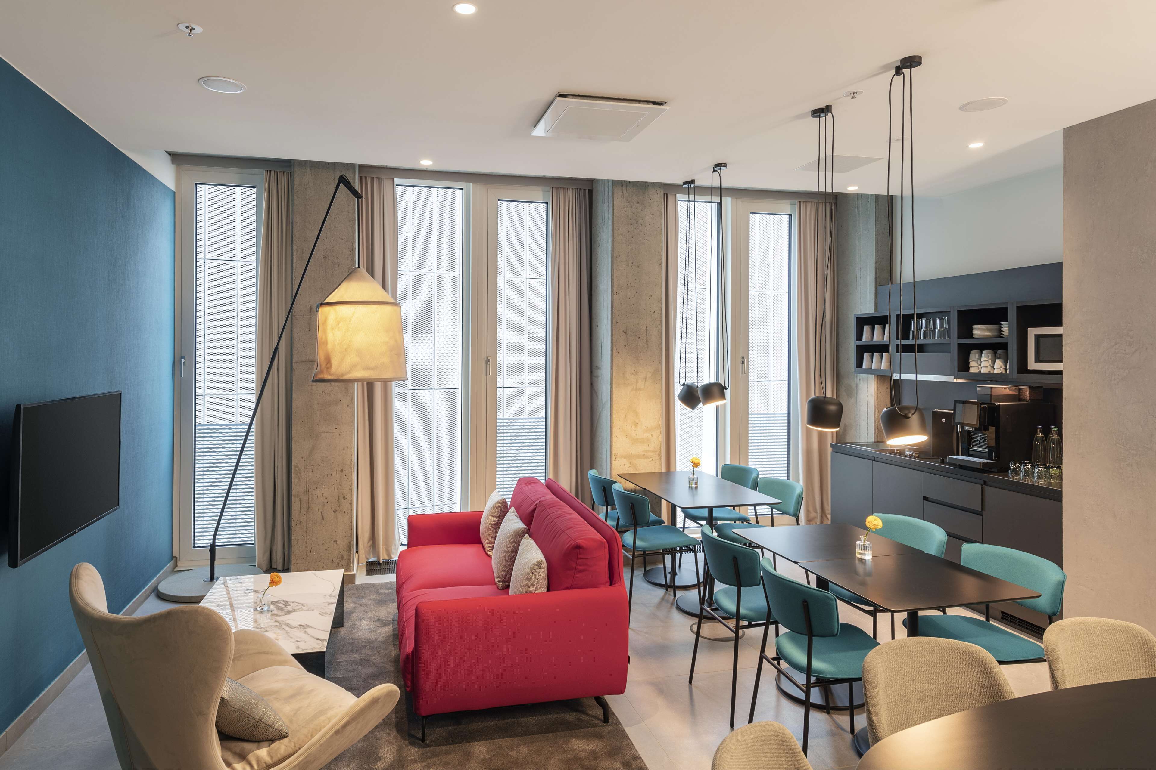 A modern living room with a red sofa, teal chairs, a dining table, and a kitchen area with a microwave and shelves.