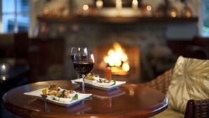 Una mesa de comedor con platos de comida y una copa de vino tinto, frente a un hogar encendido.