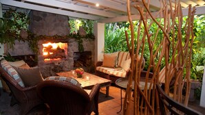 Un patio al aire libre con mobiliario de mimbre, una chimenea de piedra y abundante follaje.