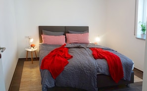 A bed with grey and red checkered pillows, a grey blanket, and red throw pillows. There is a small bedside table with a lamp and a vase with flowers. - Spacious vacation apartment with terrace & 3 bedrooms on the Swabian Alb (Blaubeuren)