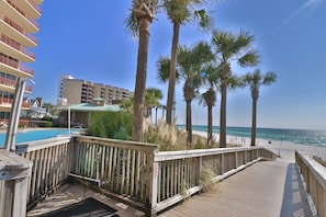 A wooden deck leading to a beach with palm trees, a pool, and a hotel building in the background.