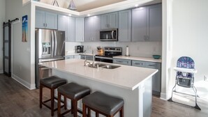 A modern kitchen with grey cabinets, stainless steel appliances, a large island with bar stools, and a small seating area. - Blue Haven – Waterfront Home w/ Heated Pool & Private Pier (Rockport)