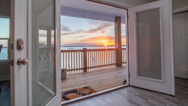 A room with a sliding glass door leading to a balcony with a wooden railing, overlooking a sunset over a body of water. - Blue Haven – Waterfront Home w/ Heated Pool & Private Pier (Rockport)