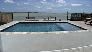 A rectangular swimming pool with a wooden deck, benches, and potted plants. - Blue Haven – Waterfront Home w/ Heated Pool & Private Pier (Rockport)