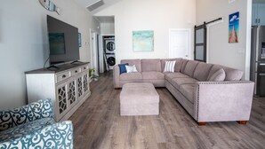 A modern living room with a sectional sofa, a TV, a dining area, and a kitchen with a refrigerator. - Blue Haven – Waterfront Home w/ Heated Pool & Private Pier (Rockport)