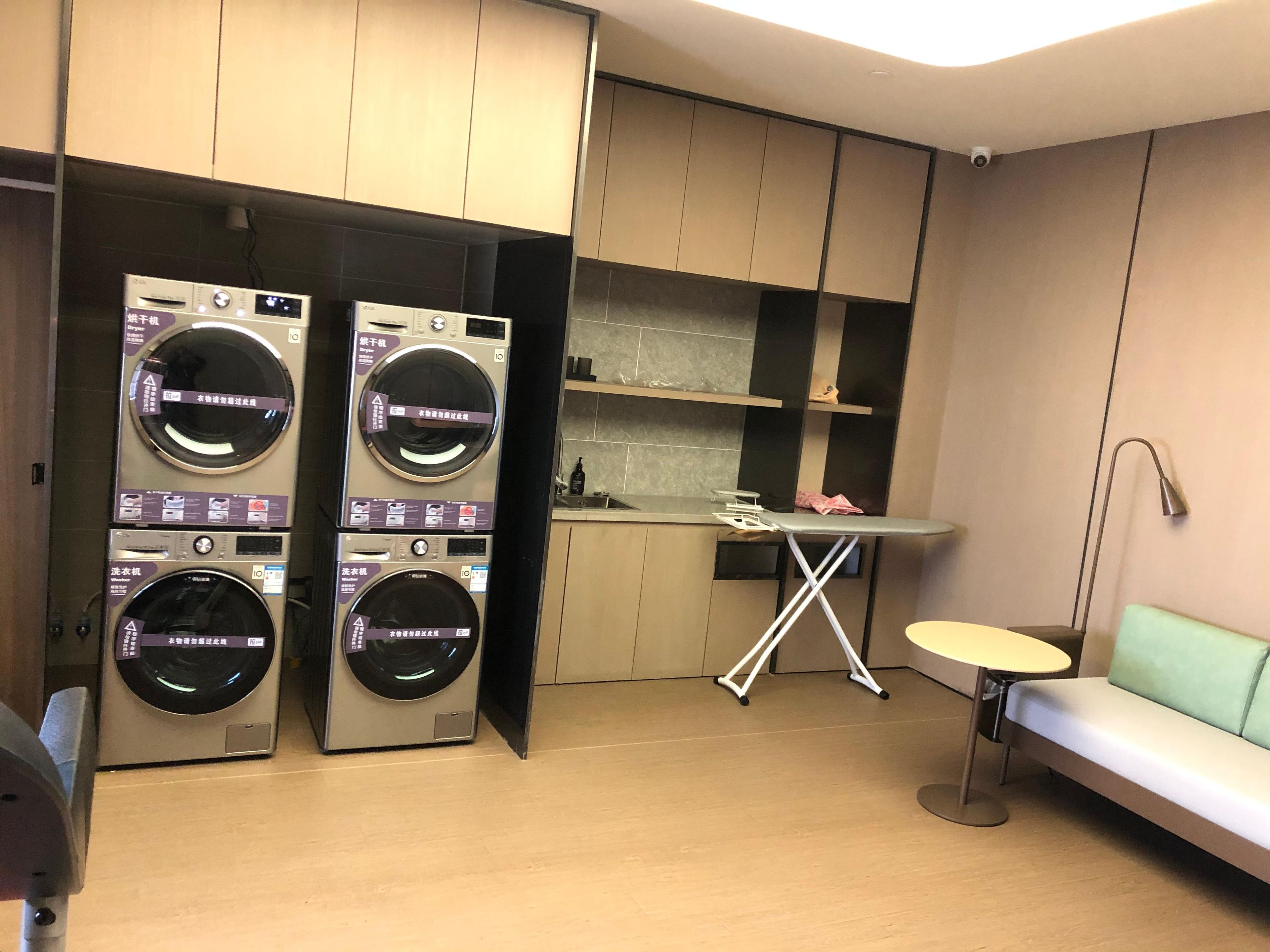 A laundry room with two stacked washing machines, a small ironing station, and a seating area.