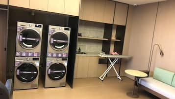 A laundry room with two stacked washing machines, a small ironing station, and a seating area.