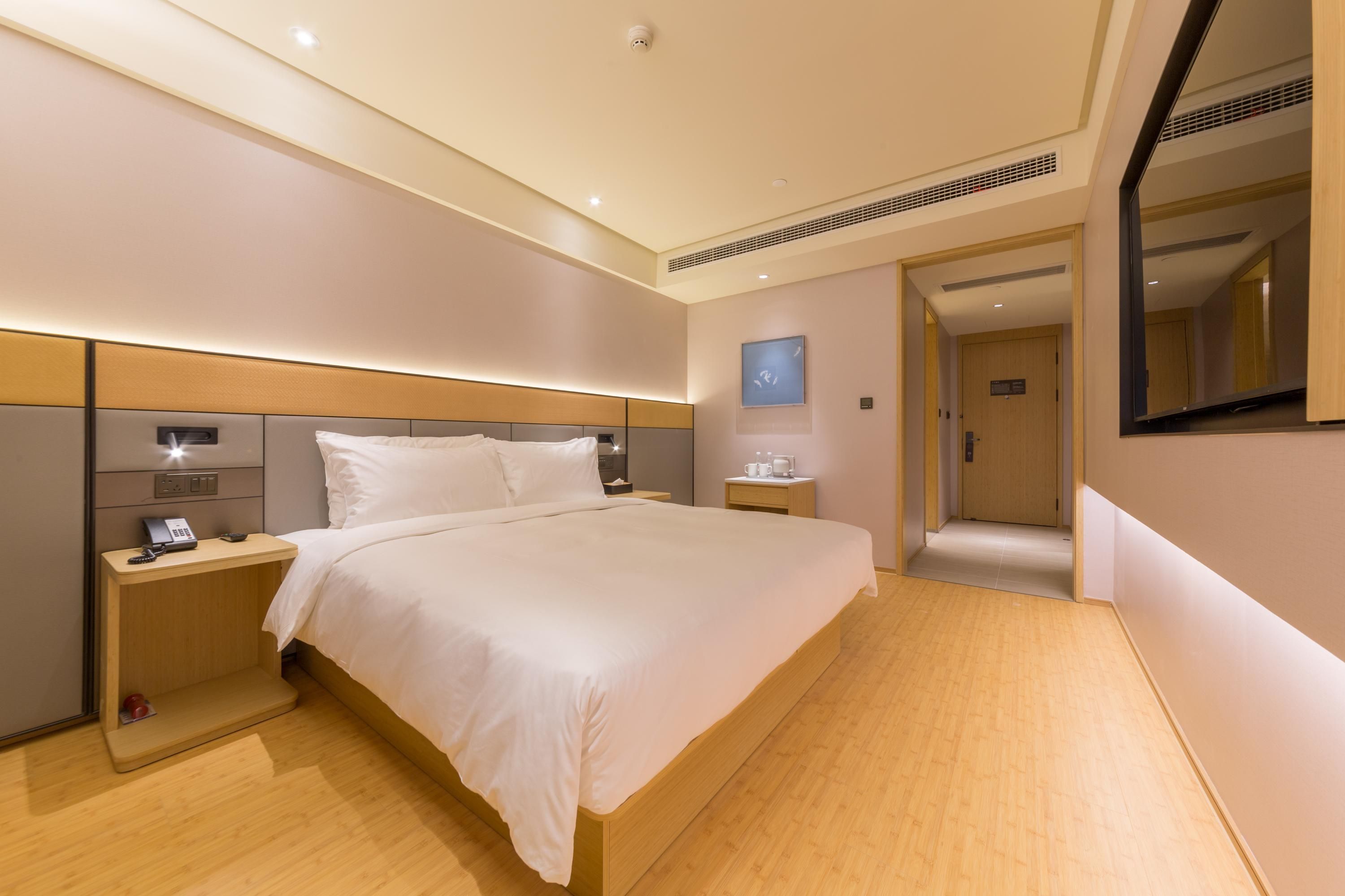 A modern hotel room with a large bed, bedside tables, a nightstand, a wall-mounted clock, and a door leading to another room.