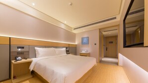 A modern hotel room with a large bed, bedside tables, a nightstand, a wall-mounted clock, and a door leading to another room.