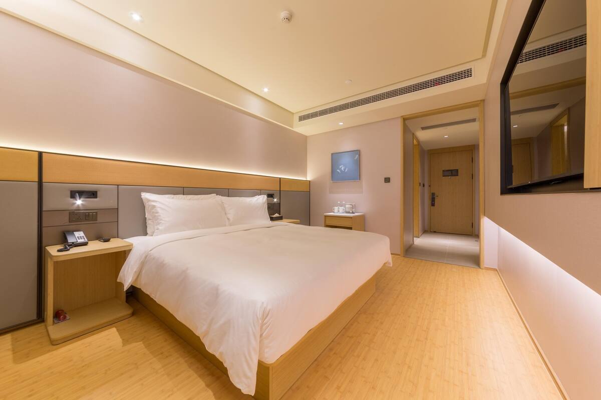 Una habitación de hotel moderna con una cama grande, mesitas de noche, una mesita auxiliar, un reloj en la pared y una puerta que da a otra habitación.