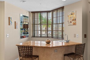 Kitchen area with central island, sink, and decorative vase, surrounded by wicker chairs and framed artwork.