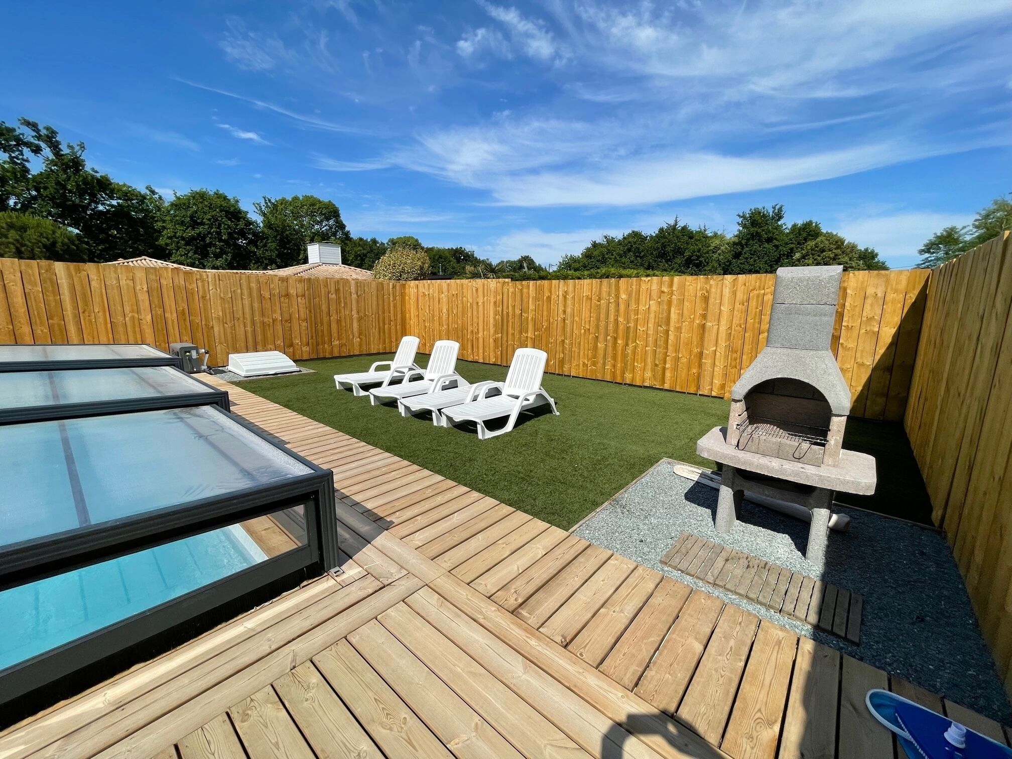 Ein Garten mit Holzterrasse, Schwimmbecken, Grill und Liegestühlen.