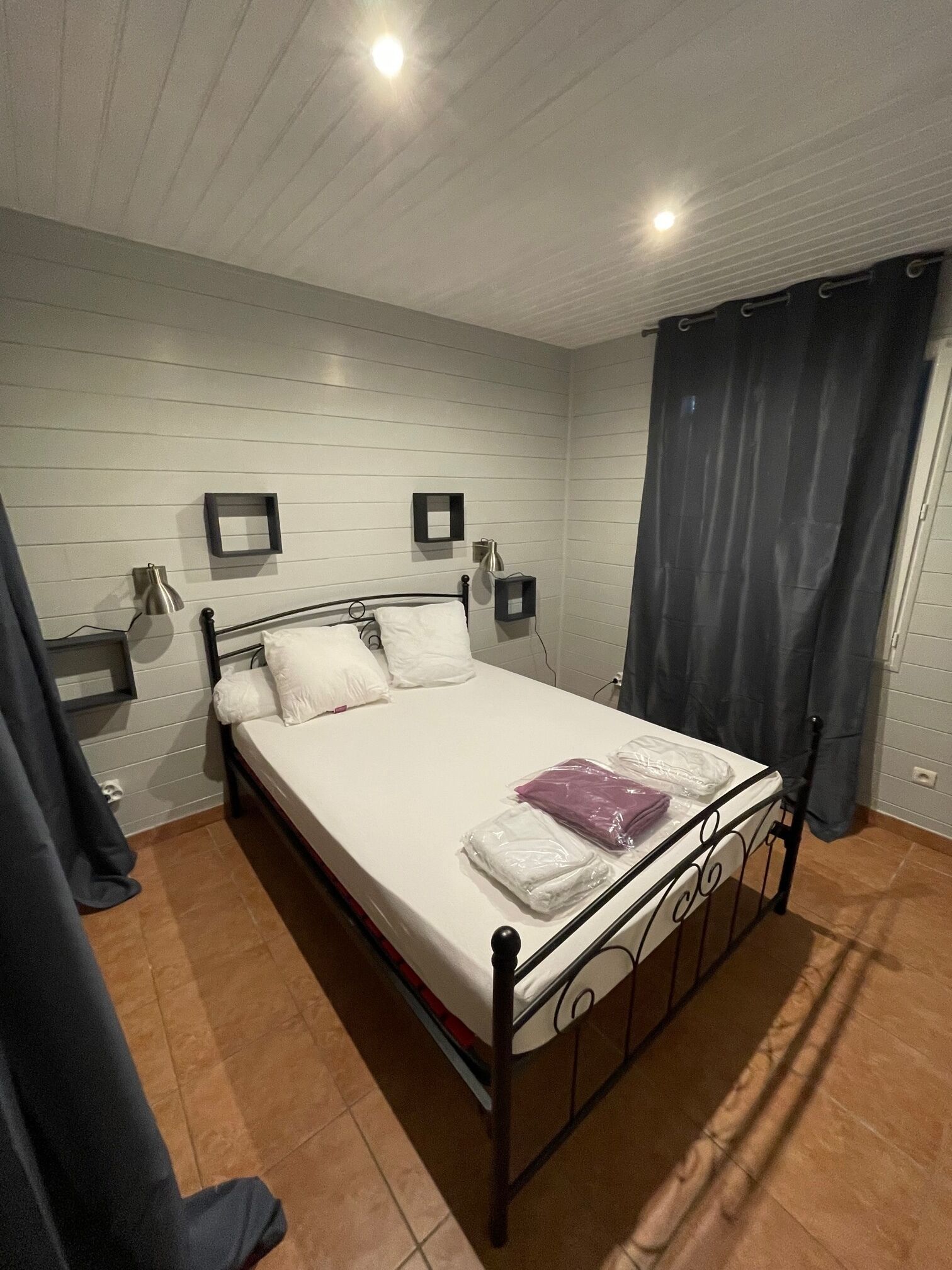 Une chambre avec un lit simple, un cadre en métal, du linge de lit blanc, une table de chevet avec une lampe et un rideau foncé.