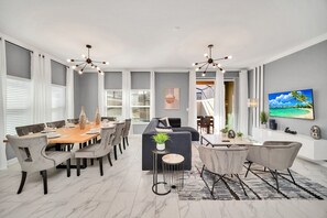 A modern living room with a dining area, grey walls, and a large window. - Luxe 5BR Townhouse w/ Pool & Mickey-Themed Room (Kissimmee)
