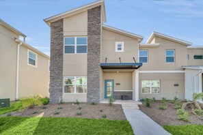 A modern two-story residential building with a stone accent wall, a front door, and a small porch. - Luxe 5BR Townhouse w/ Pool & Mickey-Themed Room (Kissimmee)