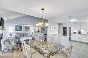 A modern living room with a glass dining table, a sofa, and a kitchen area with white cabinets. - Coastal Villa Golf Views, Pet Friendly Hilton Head (Hilton Head Island)