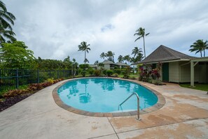 A swimming pool with a metal ladder, surrounded by palm trees and a covered area with a tiled roof.