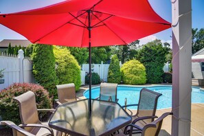 A glass-top table with chairs and a red umbrella in a backyard with a swimming pool.
