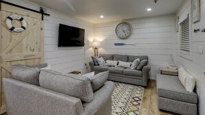 Una sala de estar acogedora con un sofá gris, un televisor de pantalla plana, un reloj de pared y un flotador colgado en la pared.