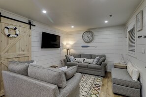 Ein gemütliches Wohnzimmer mit einer grauen Sofagarnitur, einem Flachbildfernseher, einer Wanduhr und einem Rettungsring an der Wand.