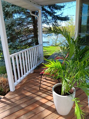 Une terrasse en bois avec une balustrade blanche, un pot de fleurs et une vue sur des arbres et une étendue d’eau.