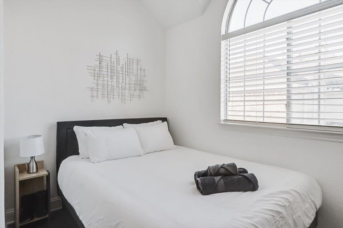 A neatly made bed with white bedding, a dark headboard, and a window with blinds.