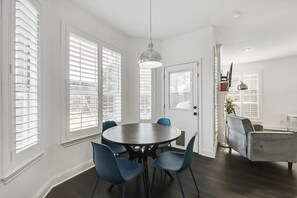 Un coin repas avec une table ronde et des chaises bleues, des murs blancs et de grandes fenêtres équipées de stores.
