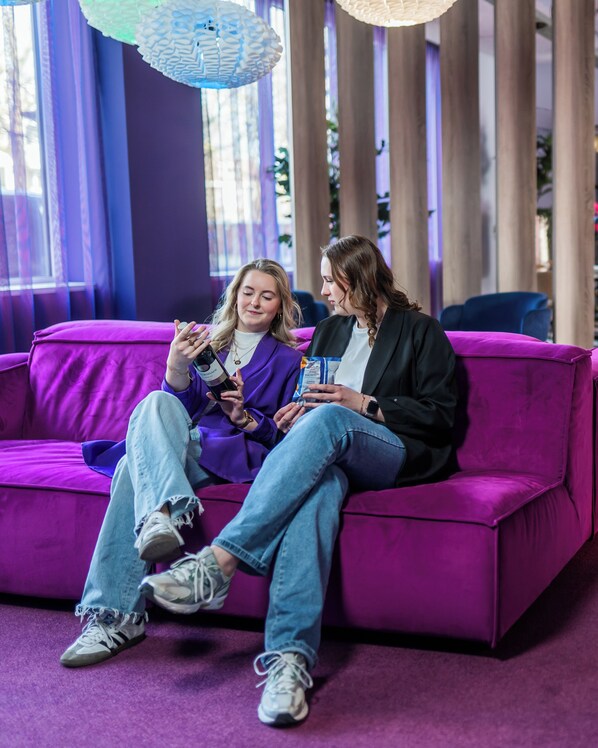 Dos personas sentadas en un sofá morado en una habitación con ventanas amplias y cortinas.