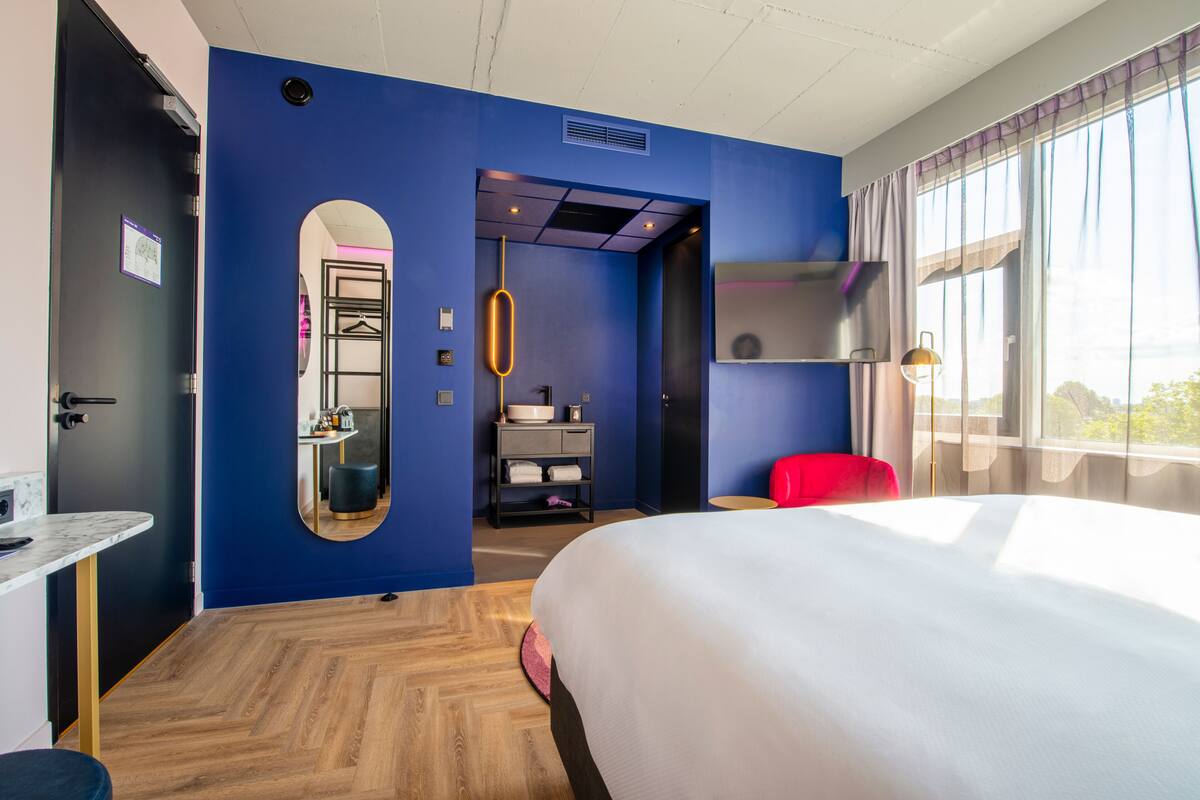 A hotel room with a large bed, a desk, a mirror, and a red chair. There is a window with curtains and a view of the outside.