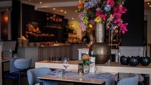 Un interior de restaurante con una zona de bar, distribución de mesas y un jarrón decorativo con flores.
